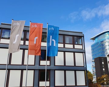 Drei Fahnen mit den Buchstaben "s", "r" und "h" vor einem modernen Gebäude mit Glasfassade und einem weiteren, traditionelleren Gebäude im Hintergrund.