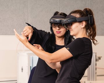 Zwei Frauen tragen Augmented-Reality-Brillen und interagieren miteinander. Eine Frau zeigt auf etwas, während die andere aufmerksam zuschaut. Ein Laptop steht auf dem Tisch in der Nähe.