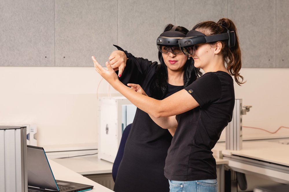 Zwei Frauen tragen Augmented-Reality-Brillen und interagieren miteinander. Eine Frau zeigt auf etwas, während die andere aufmerksam zuschaut. Ein Laptop steht auf dem Tisch in der Nähe.