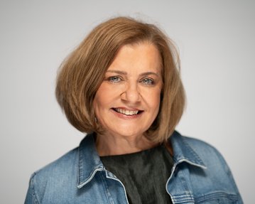 Eine lächelnde Frau mit schulterlangen, braunen Haaren trägt eine Jeansjacke und ein schwarzes Oberteil. Der Hintergrund ist neutral.