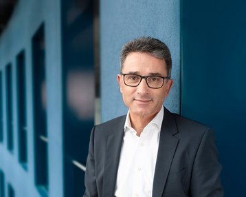 Ein Mann in einem Anzug steht an einer Wand in einem modernen Bürogebäude mit großen Fenstern und geschwungener Architektur.