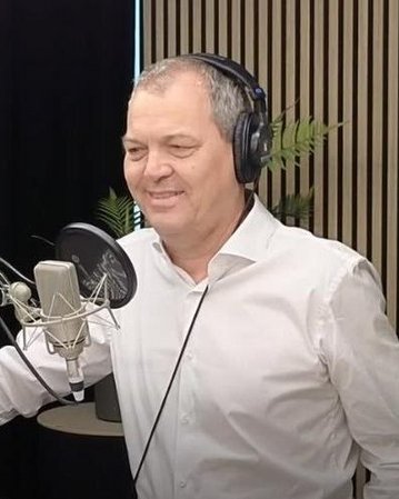 Ein Mann mit Kopfhörern steht vor einem Mikrofon in einem Studio, lächelnd, mit einem weißen Hemd und einer Tasse auf dem Tisch.