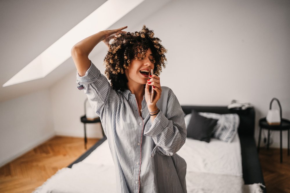 Eine Frau mit lockigem Haar trägt ein gestreiftes Hemd und singt in einem hellen Schlafzimmer. Sie hält ein Smartphone in der Hand und hat eine fröhliche Ausstrahlung.