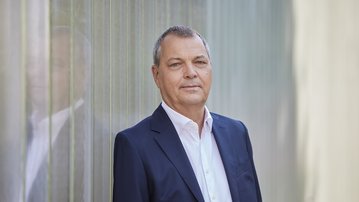 SRH Vorstandsvorsitzender Prof. Dr. Christof Hettich leitet den Konzern seit zehn Jahren Ein Mann in einem dunklen Anzug steht an einer transparenten Wand. Er hat kurzes, graues Haar und schaut direkt in die Kamera.