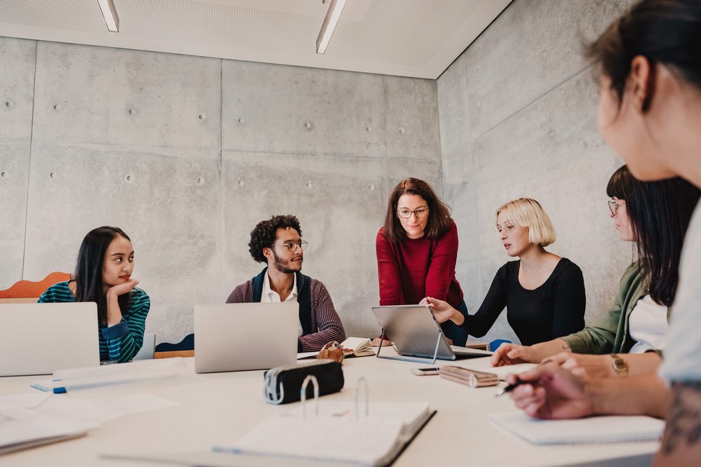 Eine Gruppe von sechs Personen sitzt an einem Tisch in einem modernen Raum. Eine Frau zeigt auf ein Tablet, während die anderen aufmerksam zuhören. Laptops und Notizblöcke sind auf dem Tisch verteilt.
