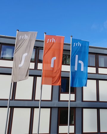 Drei Fahnen mit den Buchstaben "s", "r" und "h" stehen vor einem modernen Gebäude mit Glasfassade. Im Hintergrund ist ein weiterer moderner Bau sichtbar. Der Himmel ist teilweise bewölkt.