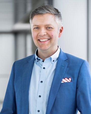 Ein Mann mit kurzen, grauen Haaren trägt einen blauen Anzug und ein hellblaues Hemd. Er lächelt freundlich und hat ein rotes Taschentuch in der Brusttasche.