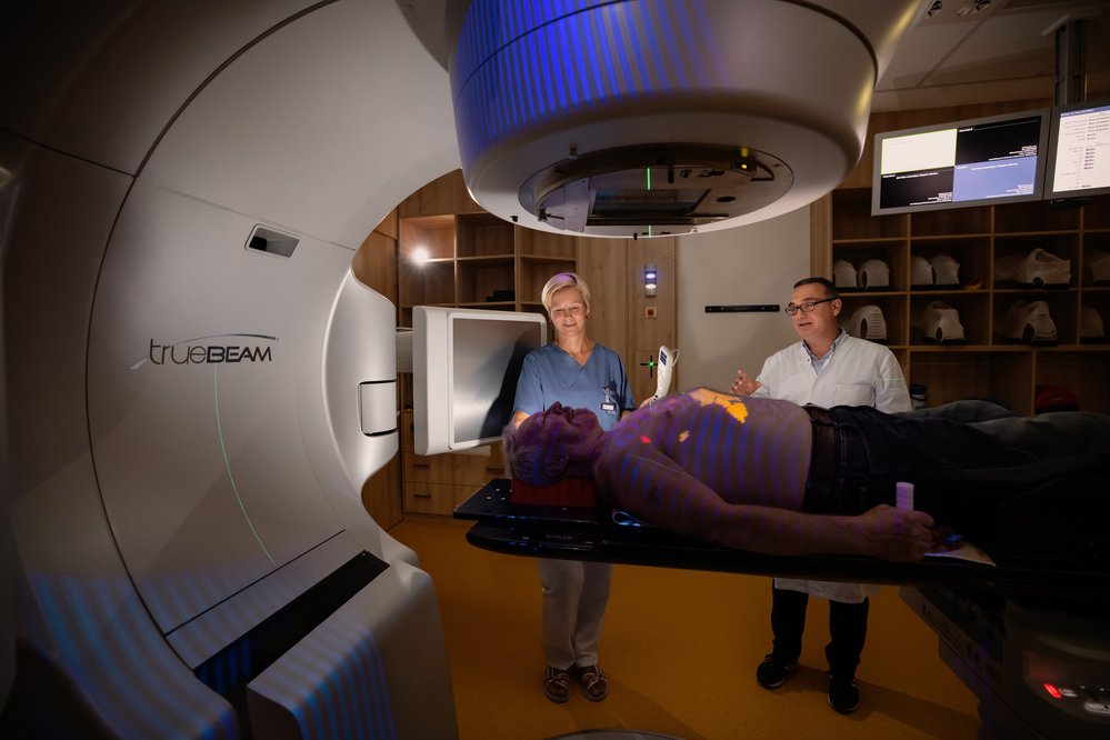 SRH Zentralklinikum Suhl 3D Sc Ein Patient liegt auf einer Behandlungsliege unter einem TrueBeam-Gerät, während zwei medizinische Fachkräfte ihn vorbereiten. Das Gerät ist für die Strahlentherapie konzipiert.