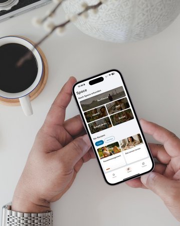 Ein Smartphone in der Hand zeigt eine App mit Kategorien wie Gesundheit, Selbst und Kurse. Im Hintergrund steht eine Tasse Kaffee.