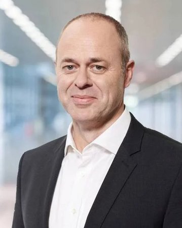 Ein Mann mit kurzen, grauen Haaren trägt einen schwarzen Anzug und ein weißes Hemd. Er steht in einem modernen Büro mit verschwommenem Hintergrund.