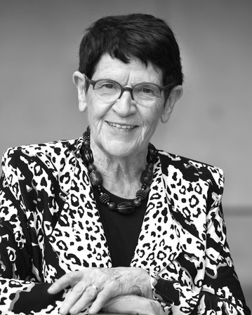 Eine ältere Frau mit Brille und einem auffälligen Leopardenmuster-Blazer lächelt und lehnt sich auf eine Tischkante.