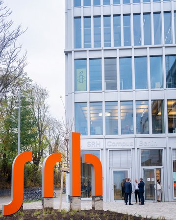 Moderne Gebäude mit großen Fenstern und einem orangefarbenen SRH-Schild im Vordergrund. Personen stehen vor dem Eingang des SRH Campus Berlin.