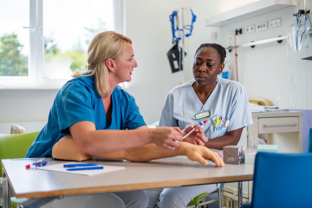 Zwei Pflegekräfte sitzen an einem Tisch und diskutieren. Eine von ihnen hält ein medizinisches Instrument in der Hand, während sie auf einen Übungsarm zeigt. Beide tragen Arbeitskleidung und scheinen sich in einem Schulungsraum zu befinden.