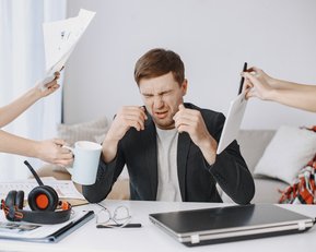 Ein Mann sitzt an einem Tisch, hält sich den Kopf und sieht gestresst aus, während Hände ihm Dokumente und eine Tasse reichen.