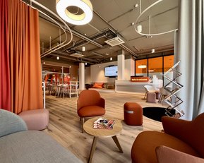 Moderne Lounge mit gemütlichen Sitzgelegenheiten, einem Tisch und einem Empfangsbereich. Ein Bildschirm zeigt "hello & welcome".