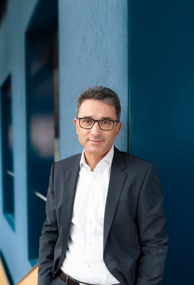 Ein Mann in einem Anzug steht an einer Wand in einem modernen Bürogebäude mit großen Fenstern und geschwungener Architektur.