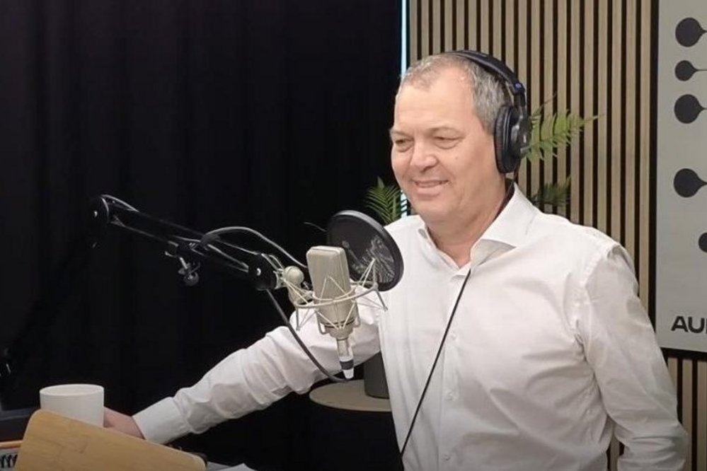 Ein Mann mit Kopfhörern steht vor einem Mikrofon in einem Studio, lächelnd, mit einem weißen Hemd und einer Tasse auf dem Tisch.