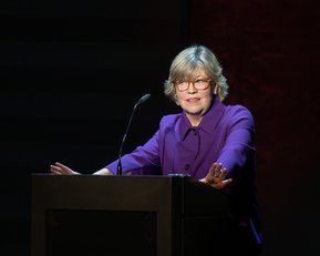 Eine Person mit blonden Haaren und Brille steht an einem Rednerpult und spricht, gekleidet in einem lila Oberteil.