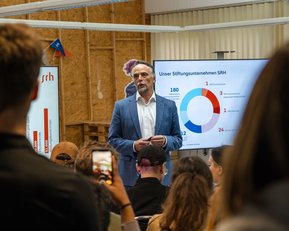 Ein Mann in einem Anzug spricht vor einem Publikum, während eine Präsentation mit Diagrammen und Statistiken im Hintergrund angezeigt wird. Die Zuschauer halten Smartphones und hören aufmerksam zu.