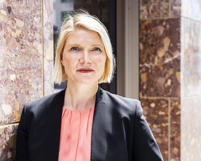 Eine Frau mit blonden, kurzen Haaren trägt ein schwarzes Blazer und ein rosa Oberteil. Sie steht an einer Wand aus Marmor und schaut direkt in die Kamera.