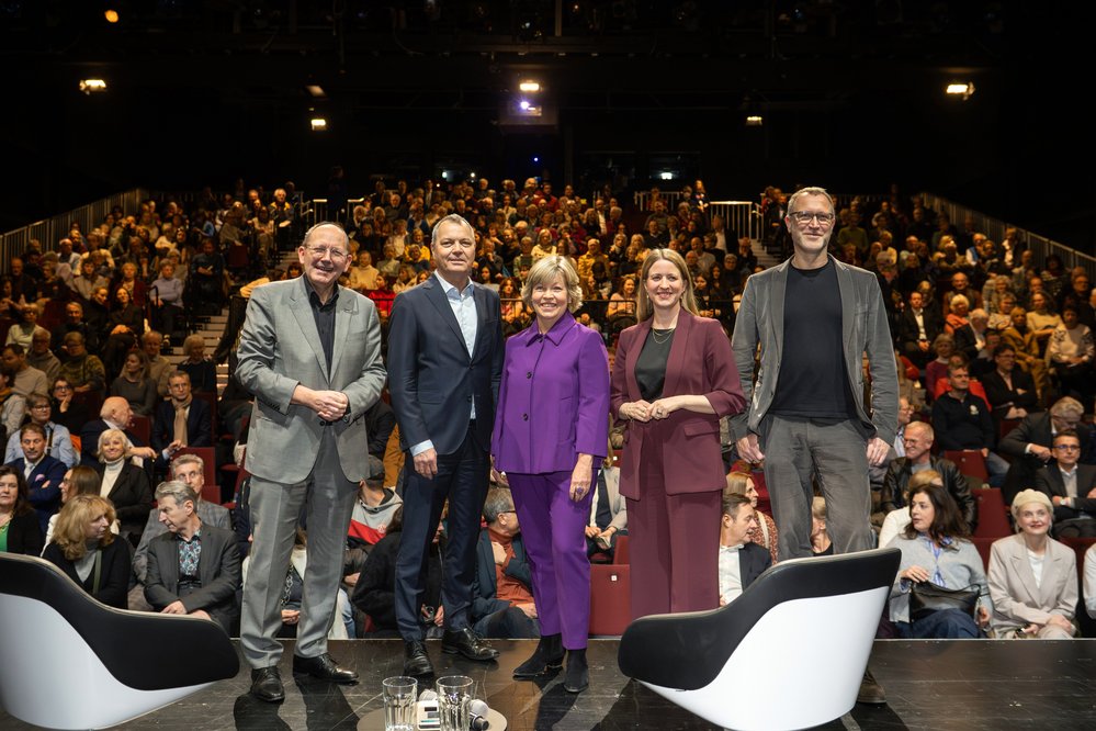 Fünf Personen stehen auf einer Bühne vor einem Publikum in einem Theater. Sie lächeln und tragen formelle Kleidung.