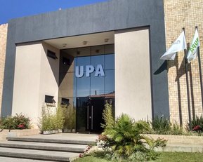 Eingang eines modernen Gebäudes mit der Aufschrift "UPA". Vor dem Gebäude befinden sich Pflanzen und Flaggen. Der Eingang ist verglast und gut beleuchtet.