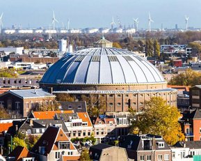 Eine große, runde Halle mit einem gewölbten Dach steht im Vordergrund, umgeben von städtischen Gebäuden und Windkraftanlagen im Hintergrund.