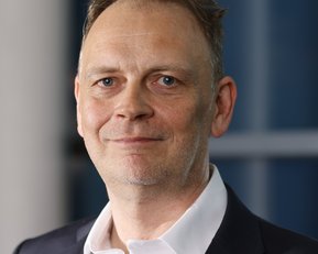 Ein Mann mit kurzen, grauen Haaren und einem leichten Bart trägt einen schwarzen Anzug und ein weißes Hemd.