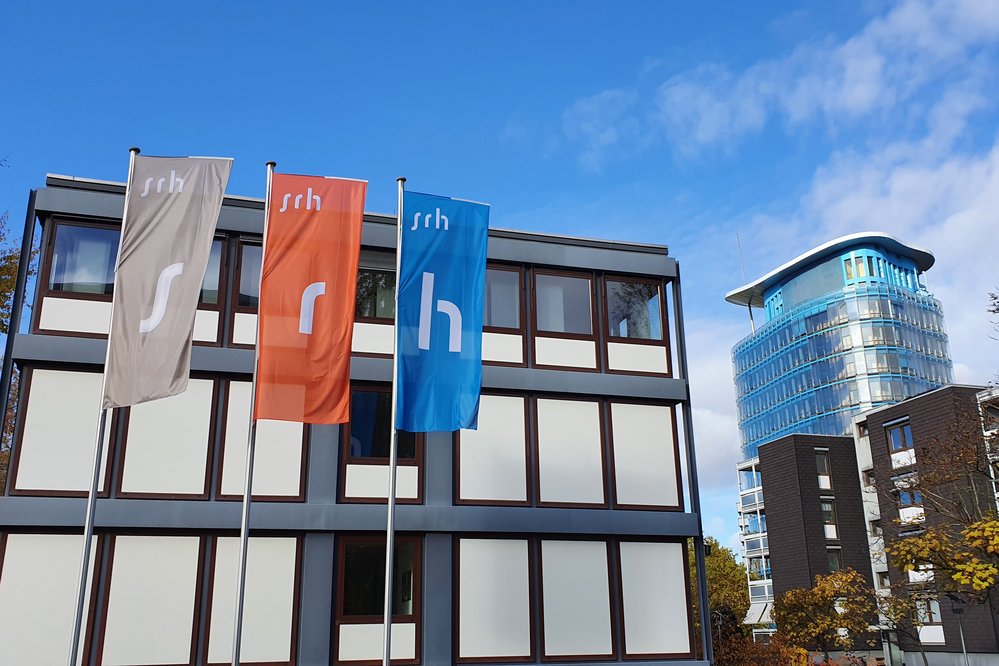 Drei Fahnen mit den Buchstaben "s", "r" und "h" stehen vor einem modernen Gebäude mit Glasfassade. Im Hintergrund ist ein weiterer moderner Bau sichtbar. Der Himmel ist teilweise bewölkt.