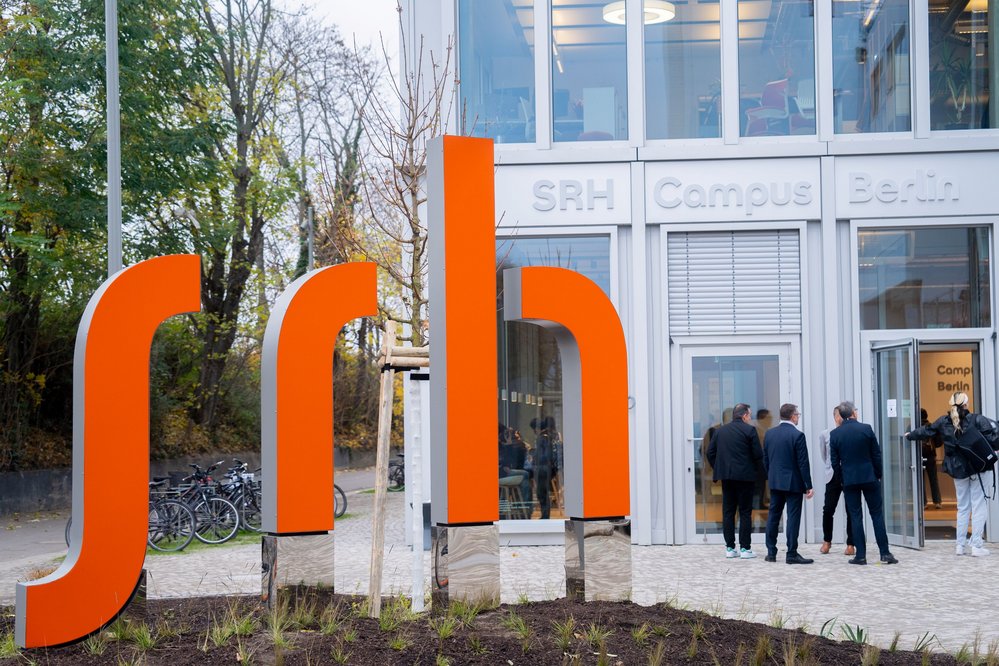 Der Eingang zum SRH Campus Berlin mit einer auffälligen orangefarbenen "srh"-Skulptur im Vordergrund. Mehrere Personen stehen vor der Tür und scheinen auf den Eintritt zu warten.