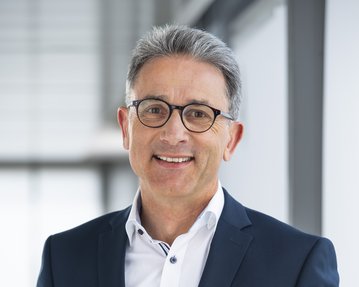 Mann in einem dunkelblauen Anzug mit Brille, lächelnd vor einem modernen Hintergrund.