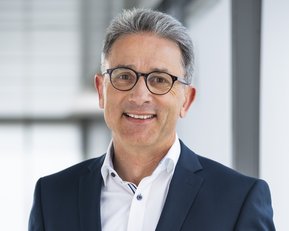 Mann in einem dunkelblauen Anzug mit Brille, lächelnd vor einem modernen Hintergrund.