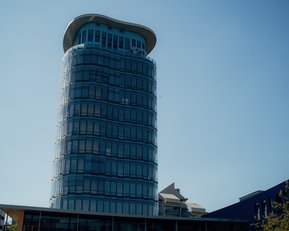 Moderne Glasfassade eines hohen Gebäudes mit abgerundetem Dach, umgeben von anderen Gebäuden und klarem Himmel.