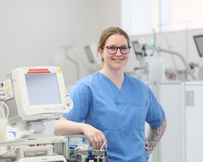 Eine Krankenschwester in blauer Arbeitskleidung steht lächelnd neben medizinischen Geräten in einem modernen Krankenhausumfeld.