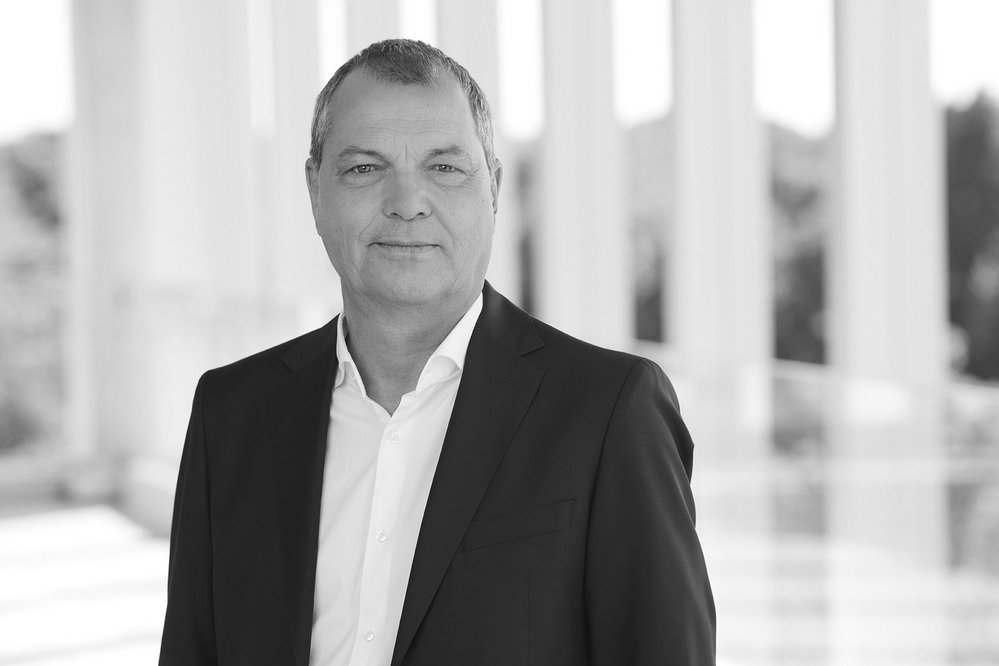 Mann in Anzug mit kurzem Haar, steht vor einer hellen, modernen Architektur. Neutraler Gesichtsausdruck, professionelles Auftreten.