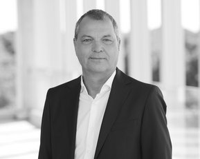 Mann in Anzug mit kurzem Haar, steht vor einer hellen, modernen Architektur. Neutraler Gesichtsausdruck, professionelles Auftreten.