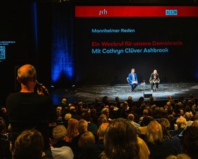 Veranstaltung mit Cathryn Clüver Ashbrook auf der Bühne, Publikum im Vordergrund, Hintergrund mit Text über Demokratie.