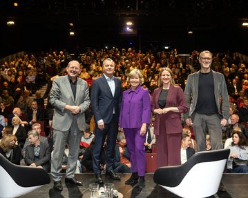 Fünf Personen stehen auf einer Bühne vor einem Publikum in einem Theater. Sie sind in formeller Kleidung und lächeln in die Kamera.