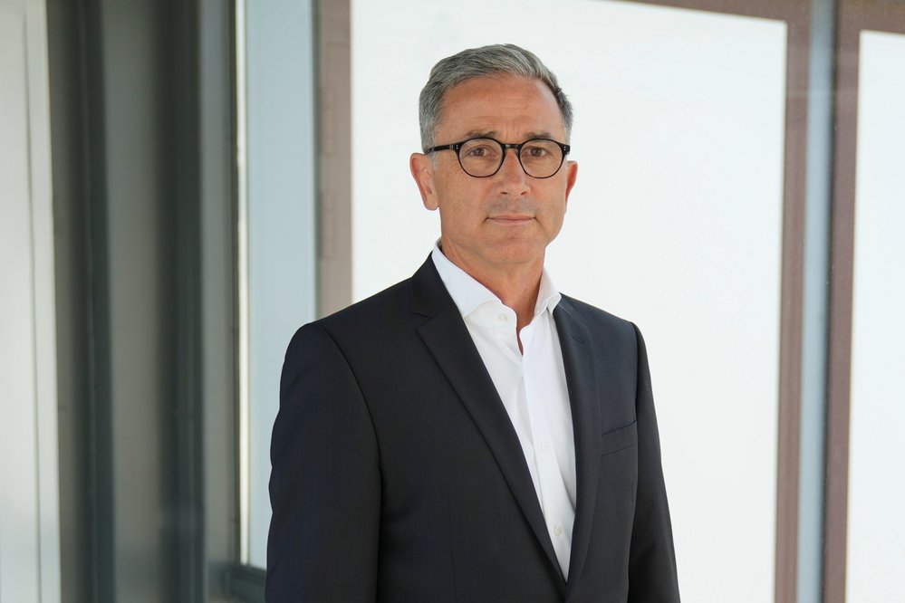 Mann in Anzug mit Brille steht vor einer modernen Glaswand. Er hat graue Haare und schaut direkt in die Kamera.