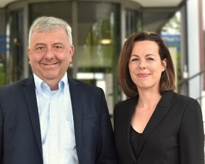 Zwei Personen stehen vor einem modernen Gebäude. Der Mann trägt einen Anzug und die Frau eine schwarze Bluse. Beide lächeln freundlich.