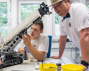 Ein Jugendlicher arbeitet an einem LEGO-Modell eines Krans, während ein Erwachsener ihm zuschaut und Anweisungen gibt.